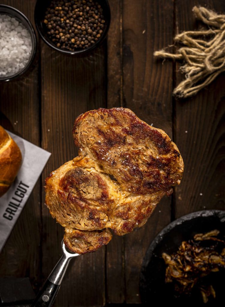 Geroestetes Fleisch auf einem Holzbrett umgeben von Gewuerzen und Kraeutern.