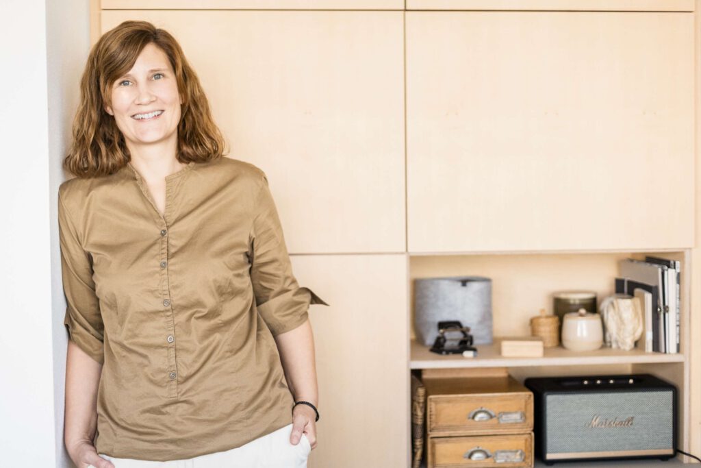 Eine Frau laechelt in einem Kuechenbereich mit Holzschraenken und Haushaltsgeraeten im Hintergrund.