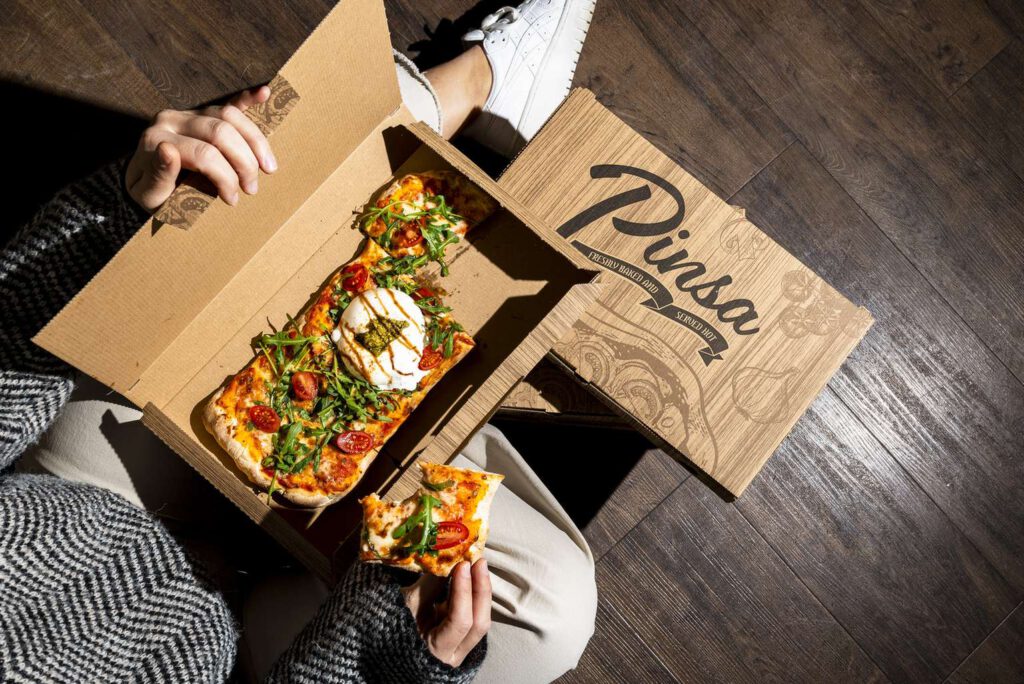 Pinsa in einem offenen Karton, gehalten von einer Person auf einem Holzboden.