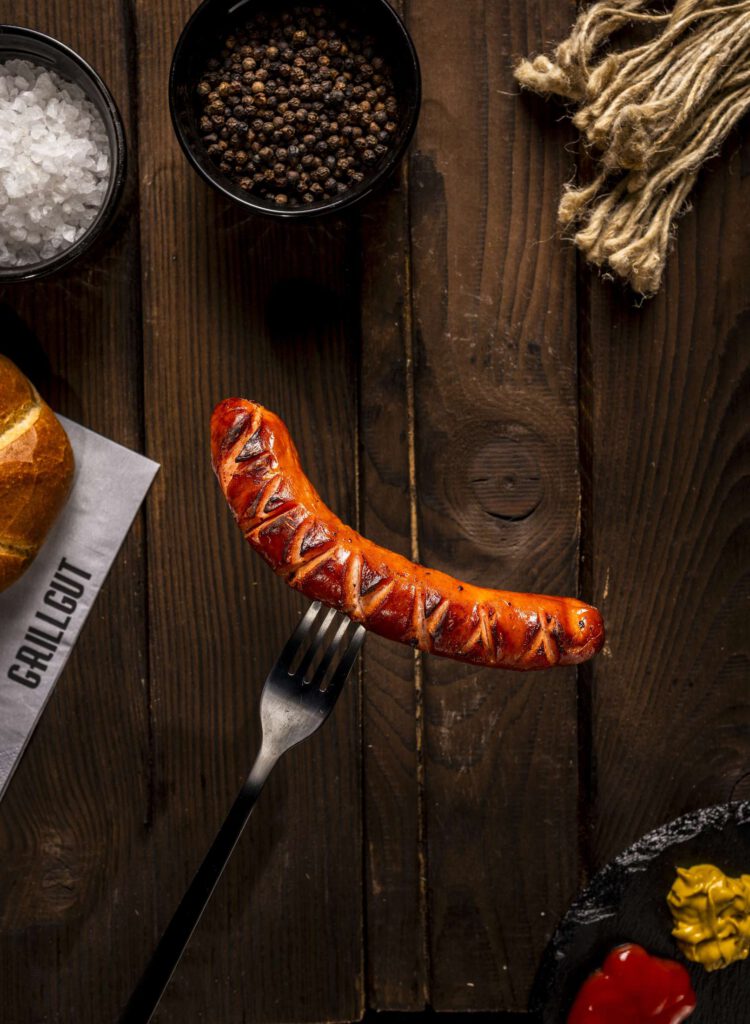 Gegrillte Wurst auf einer Gabel auf dunklem Holztisch mit Gewuerzen und Beilagen.