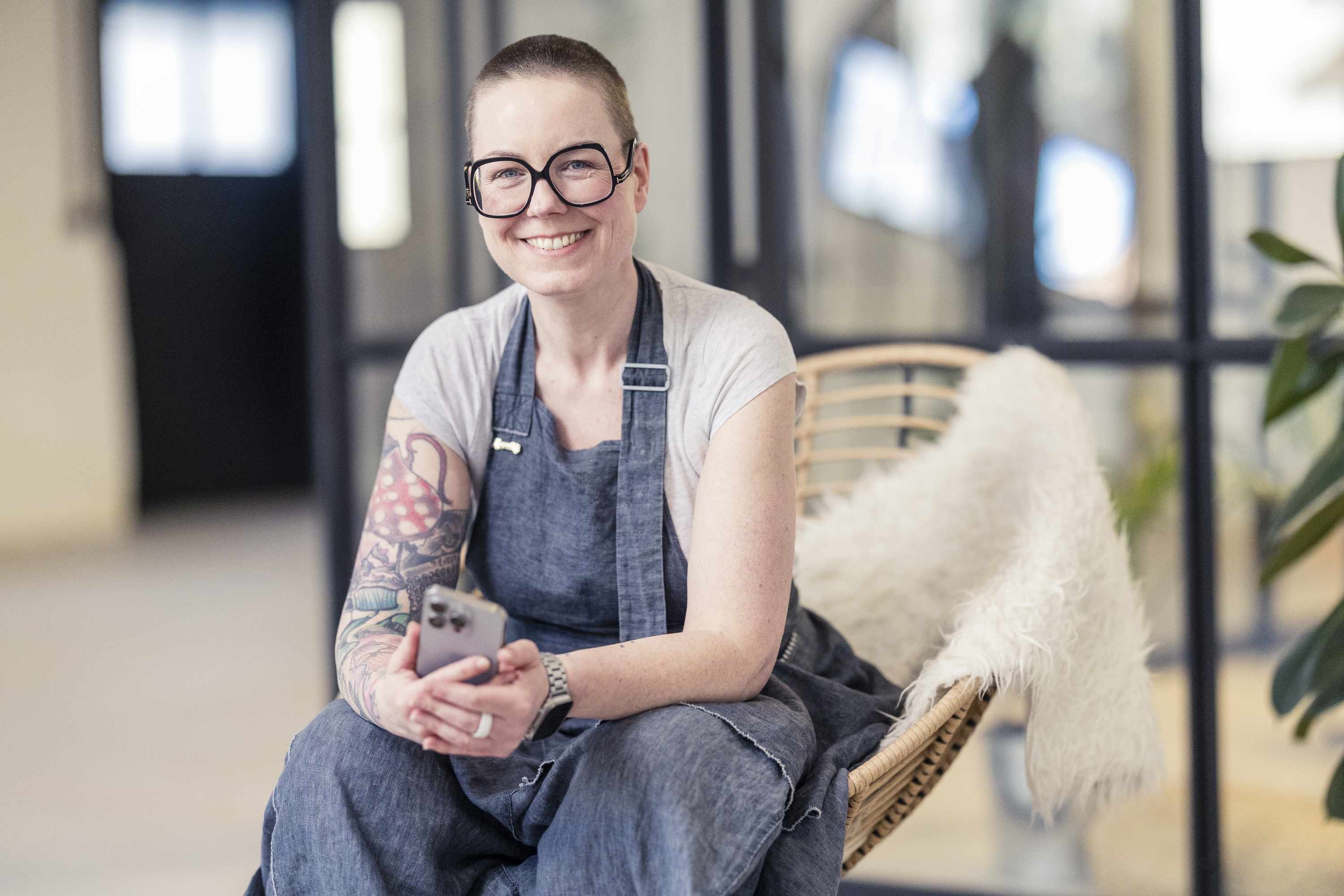Person mit Tattoos und Brille in laessiger Kleidung sitzt laechelnd auf einem Stuhl in einem modernen Buero
